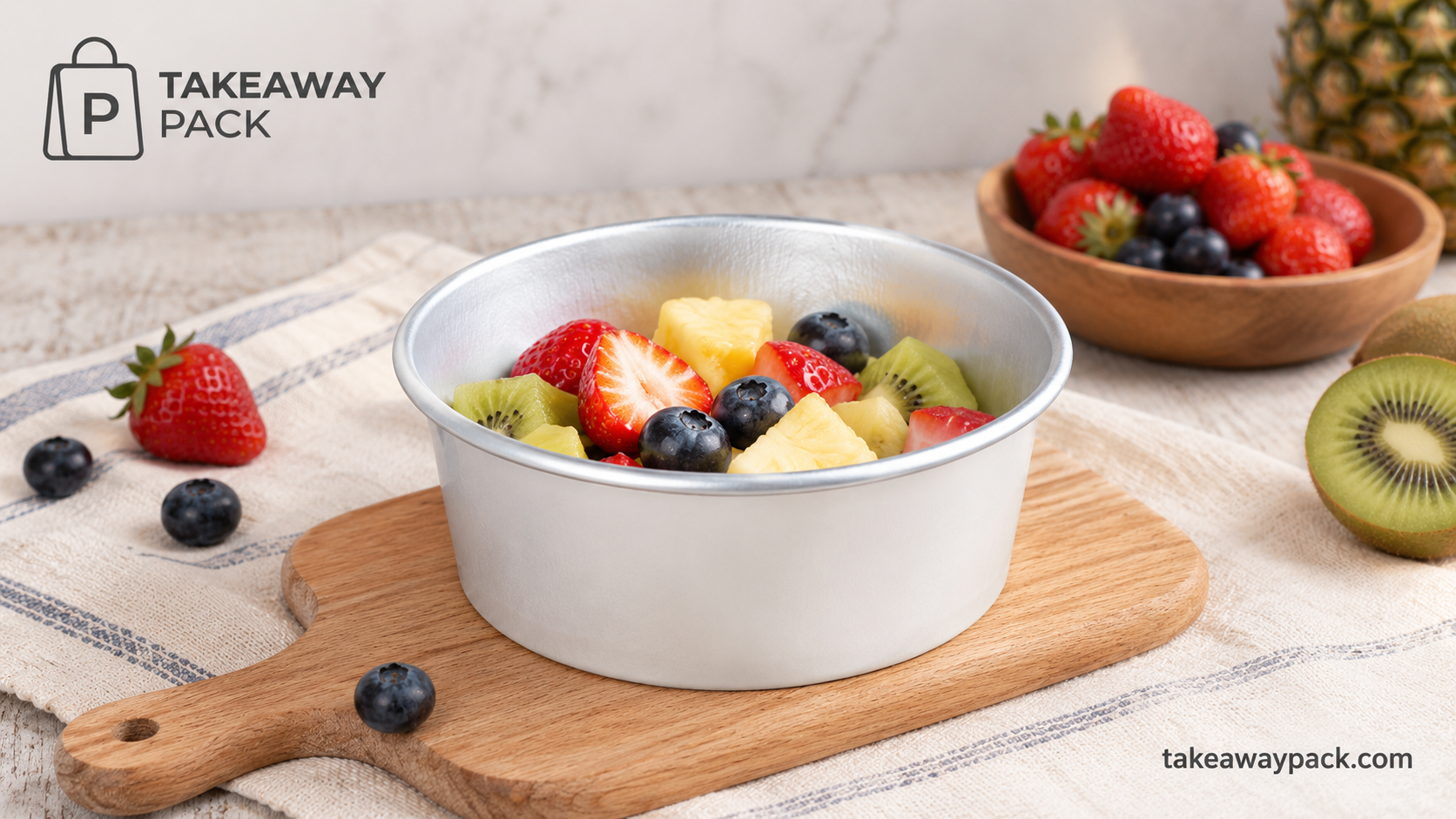 Round silver aluminium container filled with strawberries, blueberries, kiwi, and pineapple on a wooden board surrounded by more fruit.
