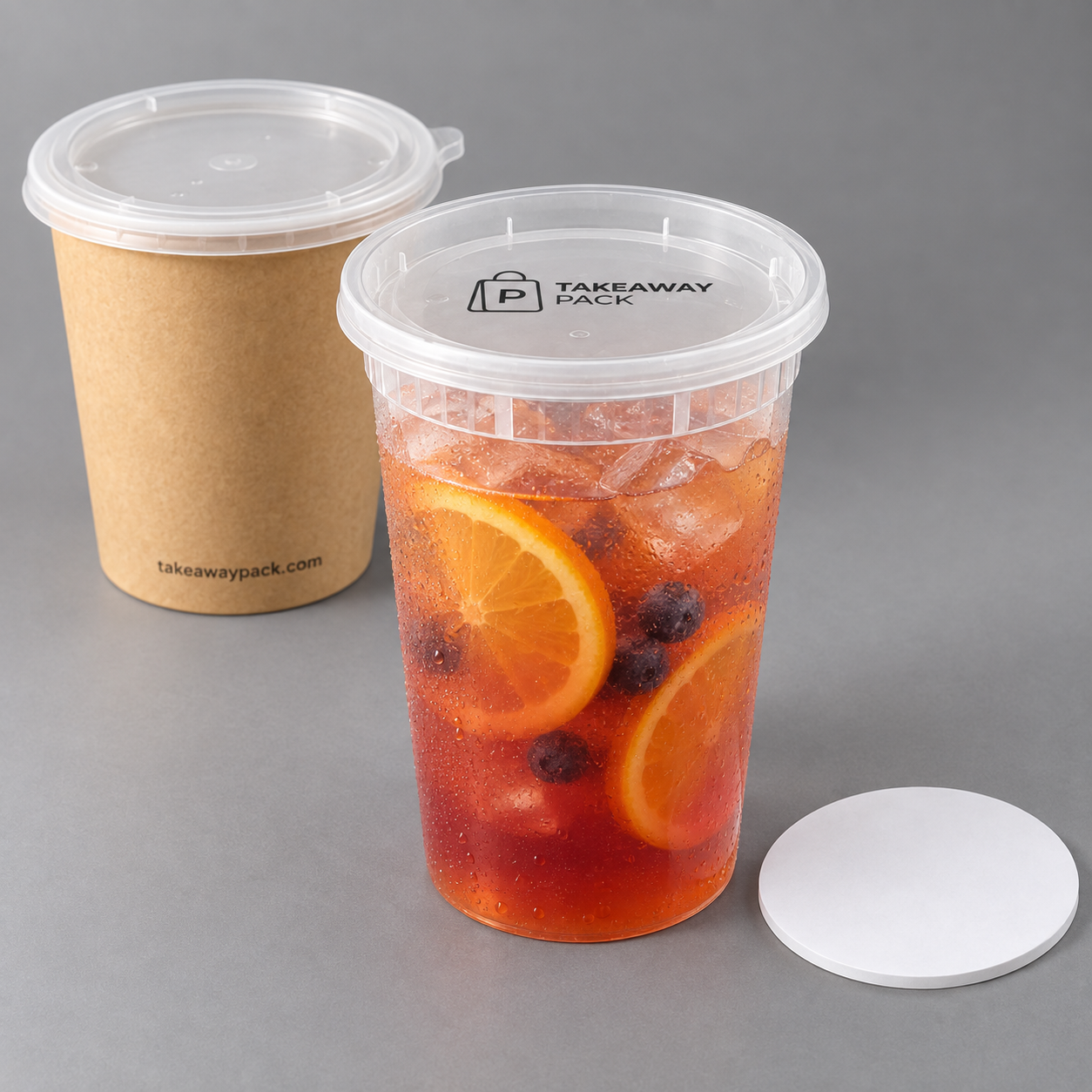 A clear cup of iced red fruit tea with orange slices and blueberries under a printed lid, a brown paper cup with a clear lid at left, and a single white lid on a grey surface.