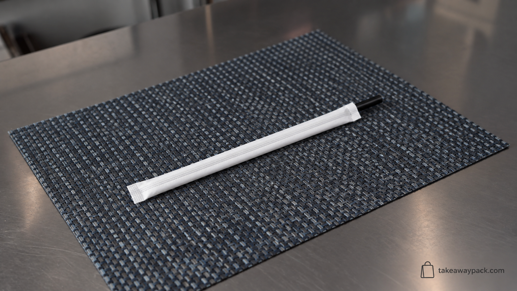 White paper-wrapped drinking straw with a glossy black tip resting on a dark grey woven placemat over a brushed metal surface.