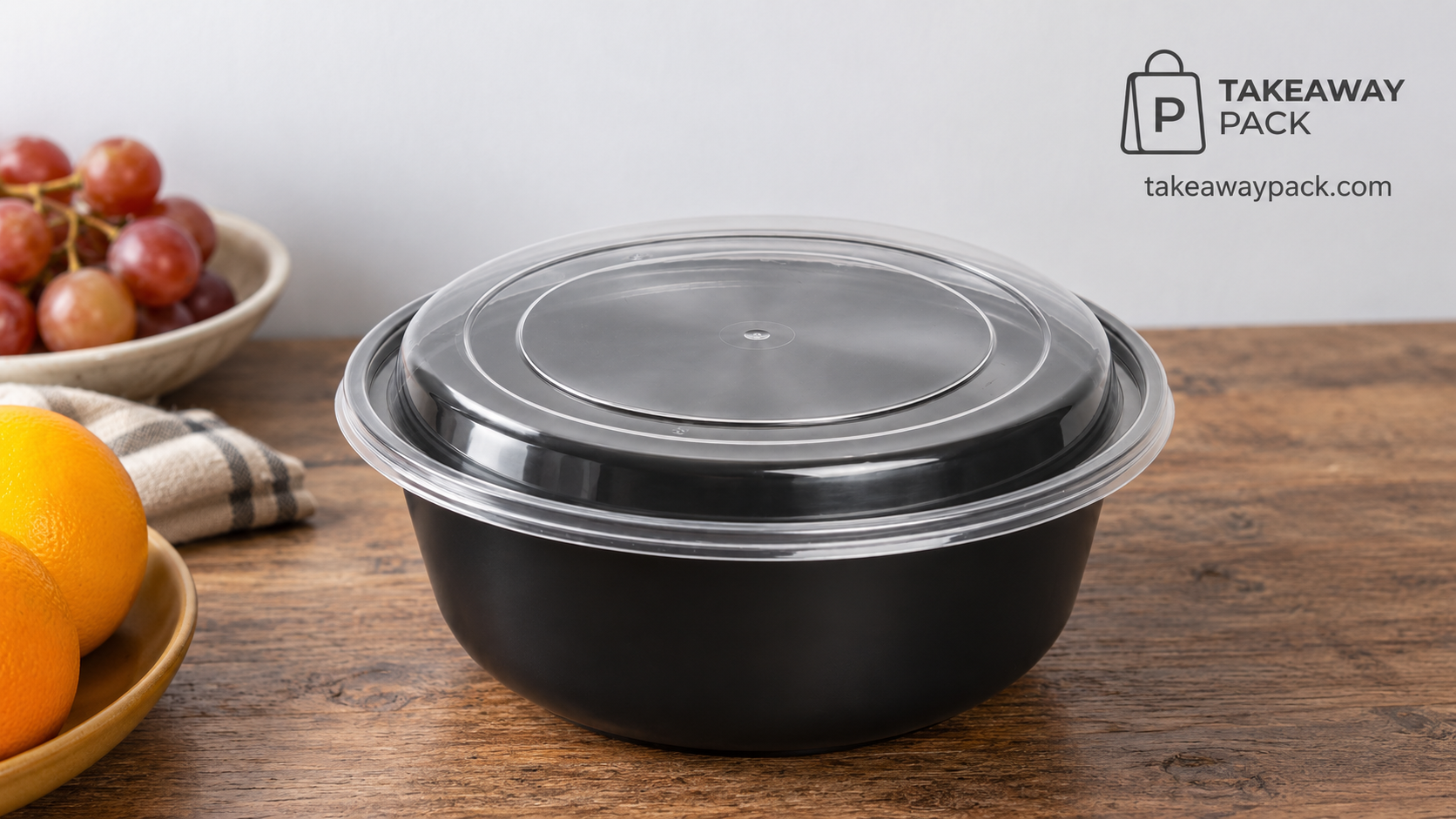 Round black plastic takeaway bowl with a clear domed lid on a wooden table beside grapes, oranges, and a striped cloth.