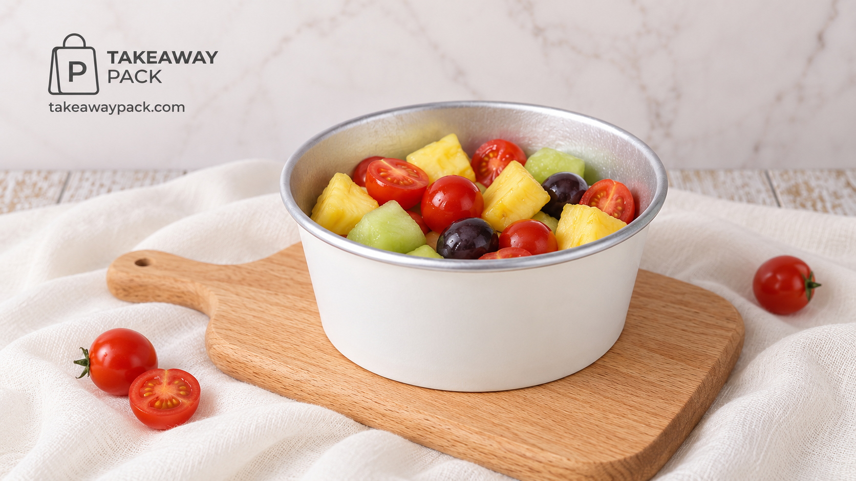 White foil-lined paper bowl of pineapple, melon, grapes, and cherry tomatoes on a wooden board over white cloth.